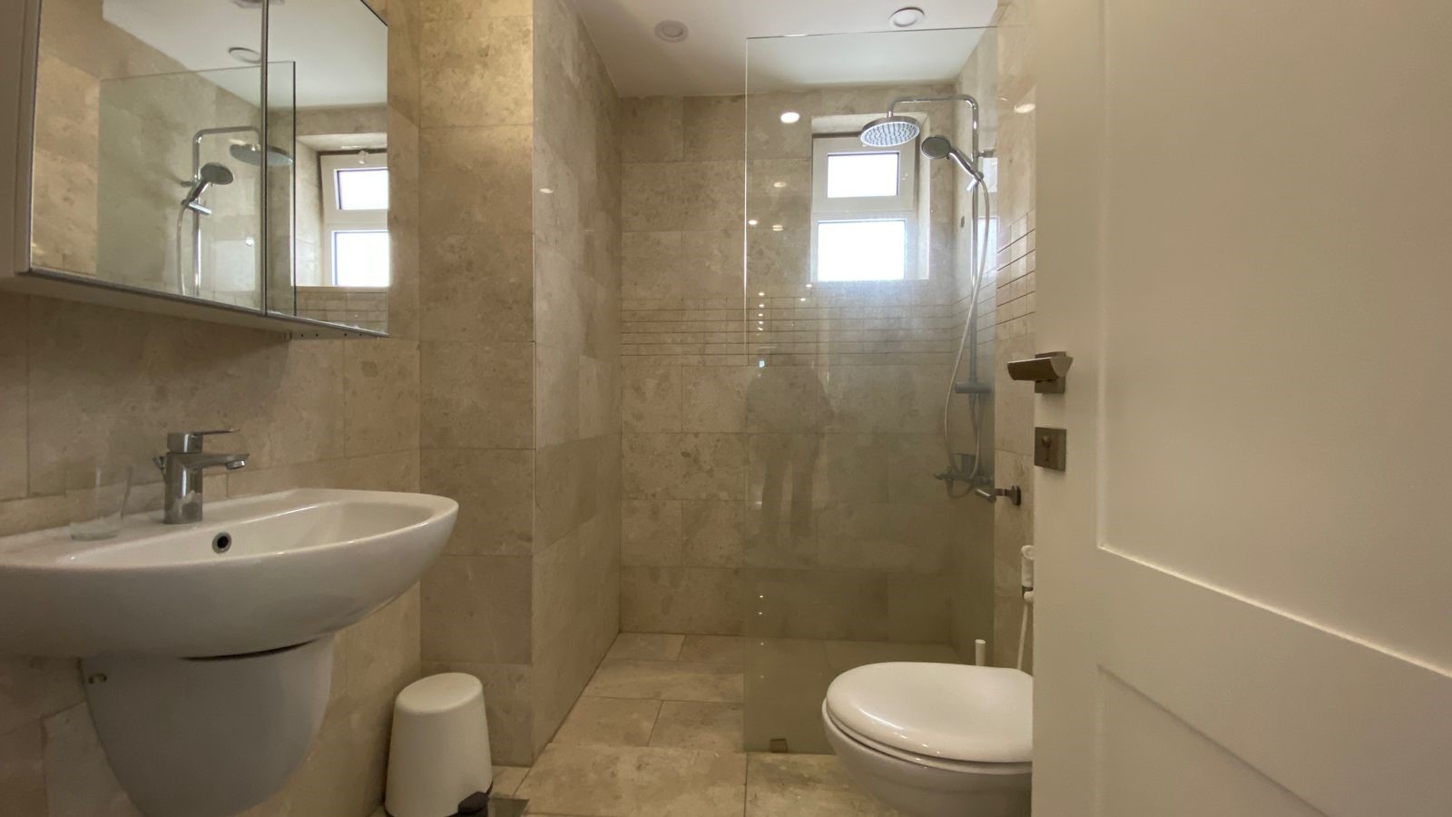 c4baf06e-5196-4b57-839d-78aa1f249c30 Beige tiled bathroom with wall mirror cabinet, white pedestal sink, glass shower enclosure, rain shower, and toilet.