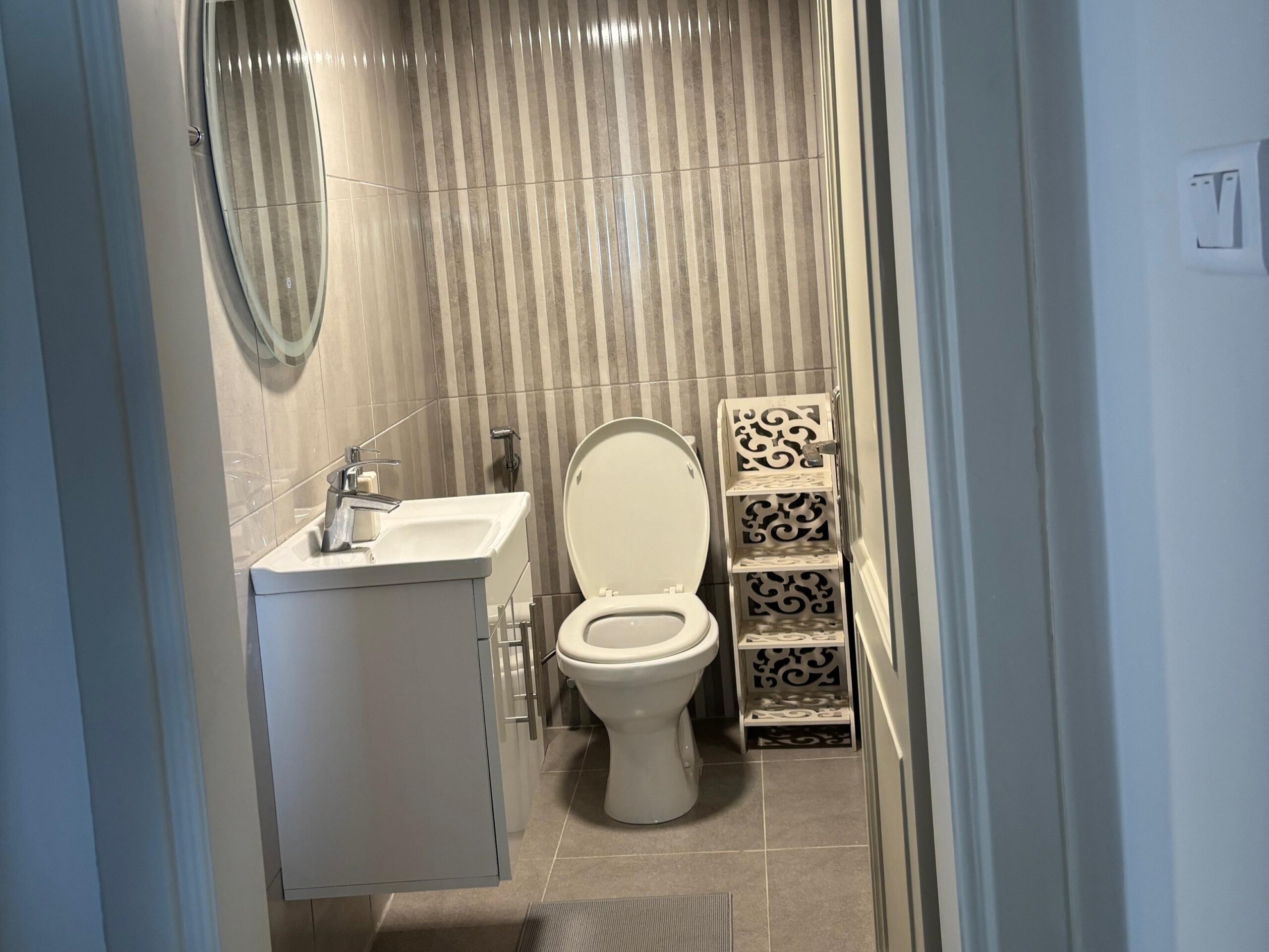 c51ce5d1-b080-4da1-a5e1-b05b95312901 Small modern bathroom with a white toilet, pedestal sink and chrome faucet, circular wall mirror, beige striped tiled wall, and a white decorative storage rack in the corner.