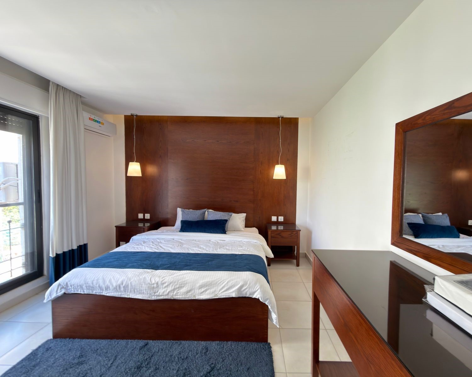 c7738e9a-0bf5-4fef-86d0-3052be2e202b Hotel bedroom with a wooden headboard, white bedding, blue accent pillows, and twin pendant lights over each nightstand.