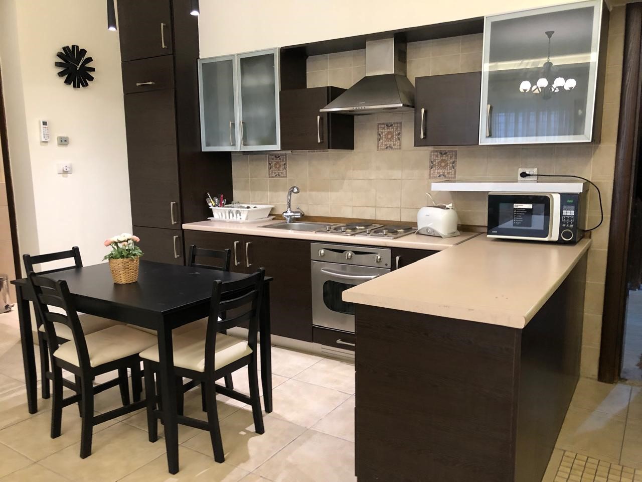 c7aa5fb4-9200-4fd6-8533-df77b86e220f Modern dark-wood kitchen with beige counters and a four-seat black dining table set, center centerpiece flowers, and cream-tile floor.