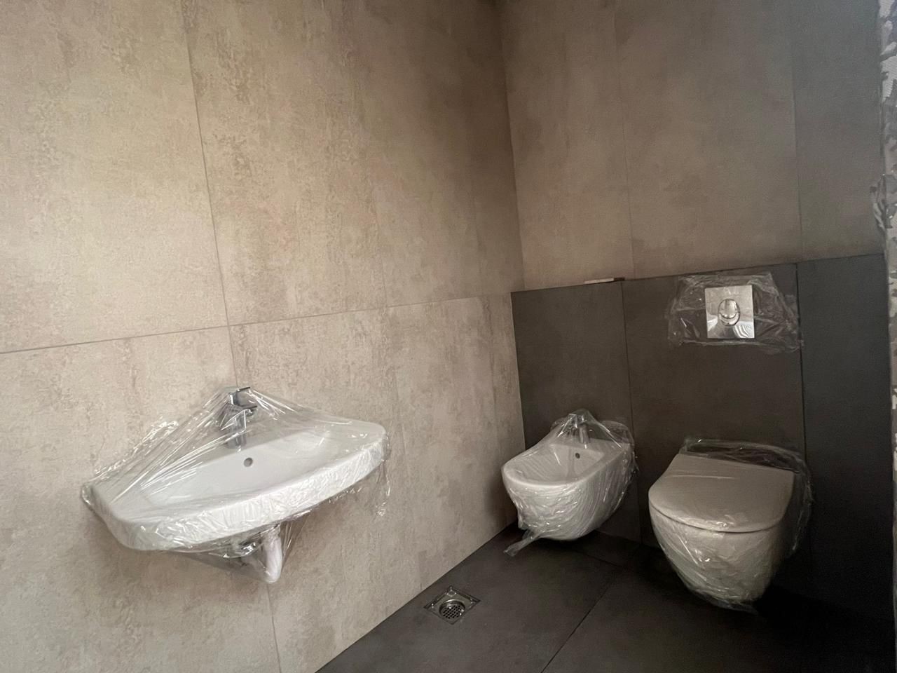 c7e4124d-3aa4-41de-bf1e-261979380ba5 New, unfinished bathroom with a wall-mounted sink, a bidet, and a wrapped toilet covered in plastic. Floor drain visible.