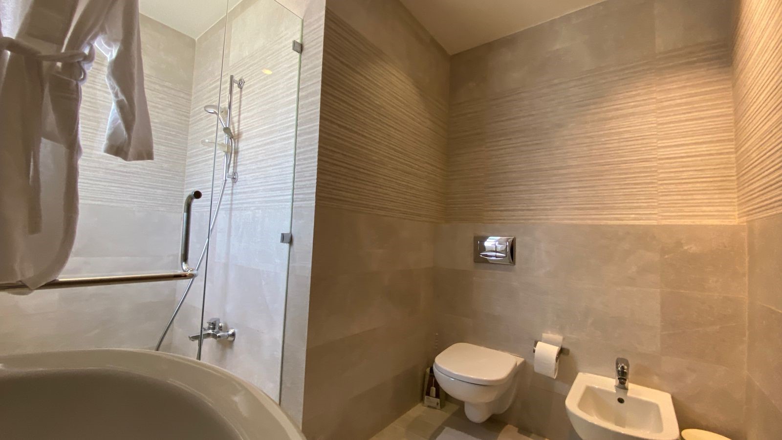 c93ec1cb-91ee-40ee-90c5-4c0de779aed9 Contemporary bathroom with a glass shower enclosure, wall-mounted toilet, and bidet, all in beige tiles.