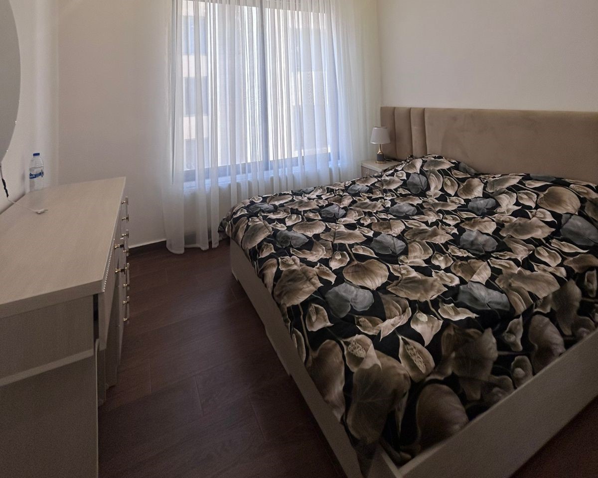 c94e745f-e9f8-41cf-913c-6e02cabf6204 Modern bedroom with a large bed covered in a dark floral duvet, a white dresser to the left, and sheer white curtains over a sunny window.