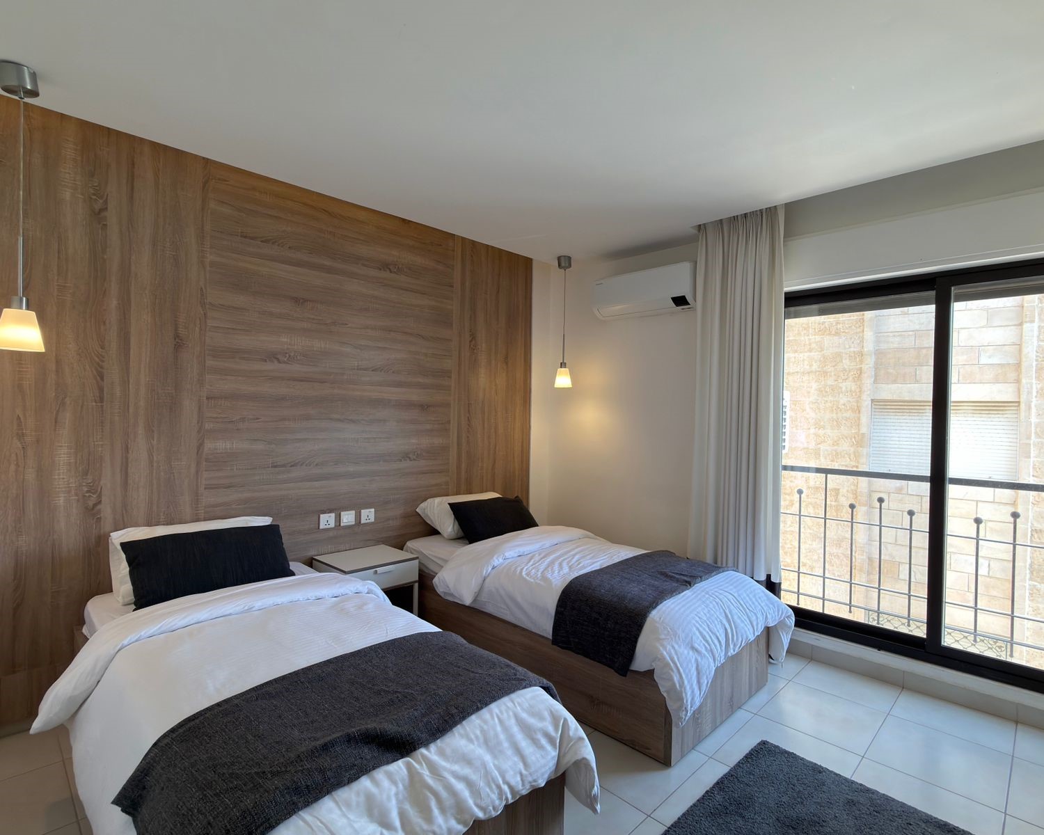 ca2ab4ea-a500-4c93-a68e-c9ba011cf469 Twin bedroom with wood panel headboard, two single beds, pendant lights, and a balcony door with white curtains.