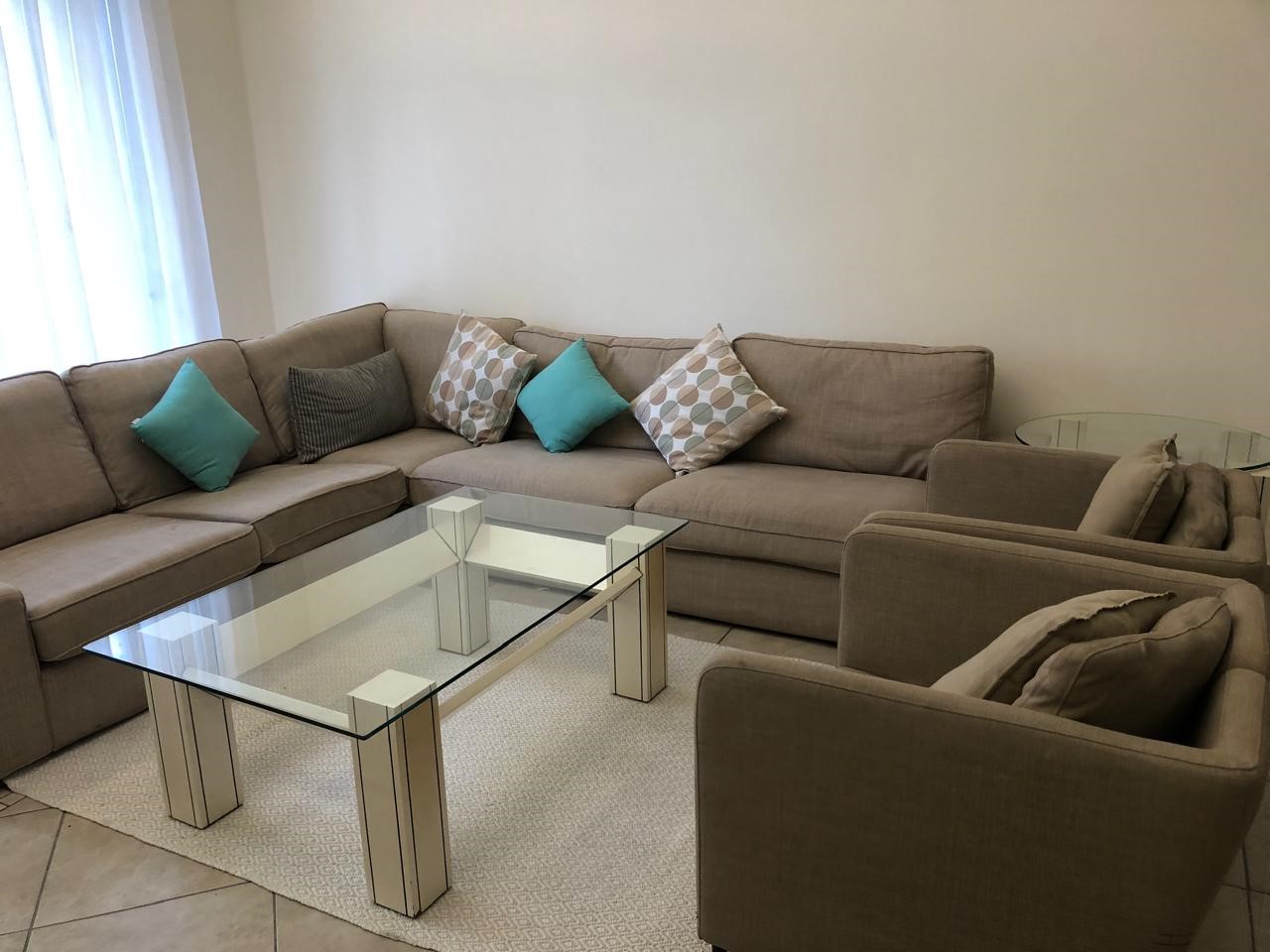 cb5264b3-05c6-449a-b86a-abdca990d983 Beige L-shaped sofa with teal and patterned cushions around a glass coffee table in a bright living room.