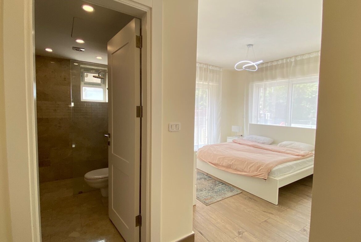 Bright bedroom with a white bed and peach duvet, large windows with sheer curtains; doorway to a tiled bathroom with a toilet visible on the left.