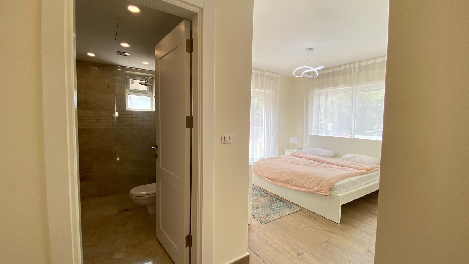 cd310088-2081-4856-a444-56c384ef68a9 Bright bedroom with a white bed and peach duvet, large windows with sheer curtains; doorway to a tiled bathroom with a toilet visible on the left.