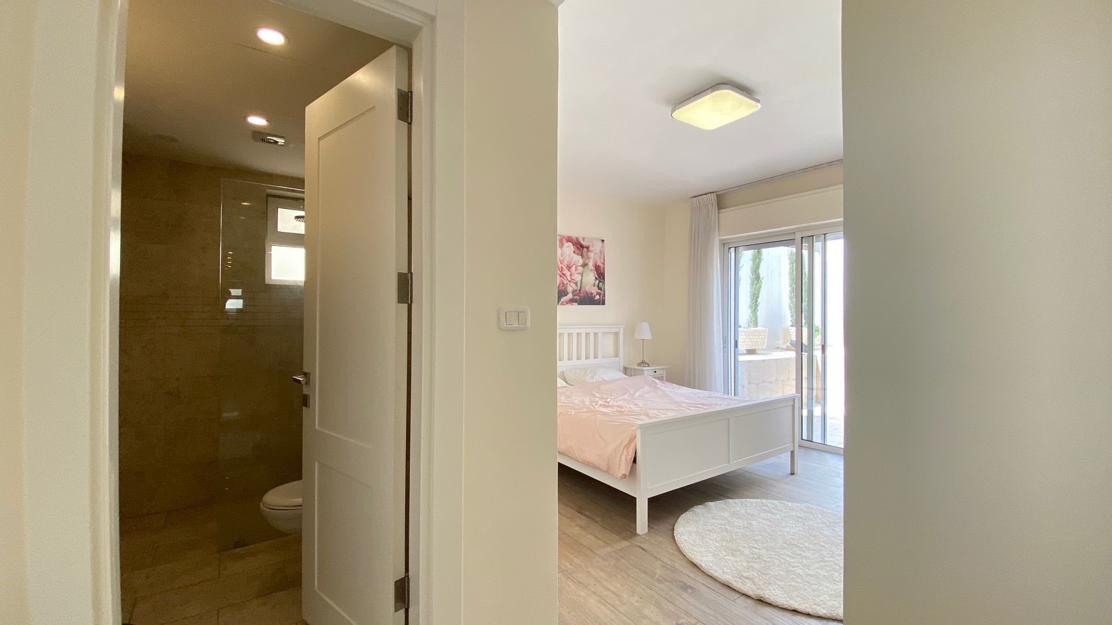 cd9fcc96-777f-4447-8317-3d277cad2d74 Bright bedroom with white bed, nightstand lamp, and a sliding glass door to a balcony; bathroom doorway visible on the left.