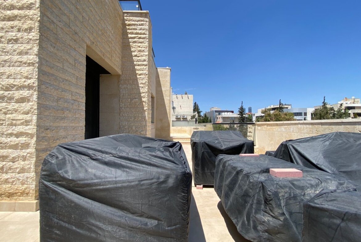 Rooftop terrace with beige stone walls and several large items covered in black tarps, against a clear blue sky.