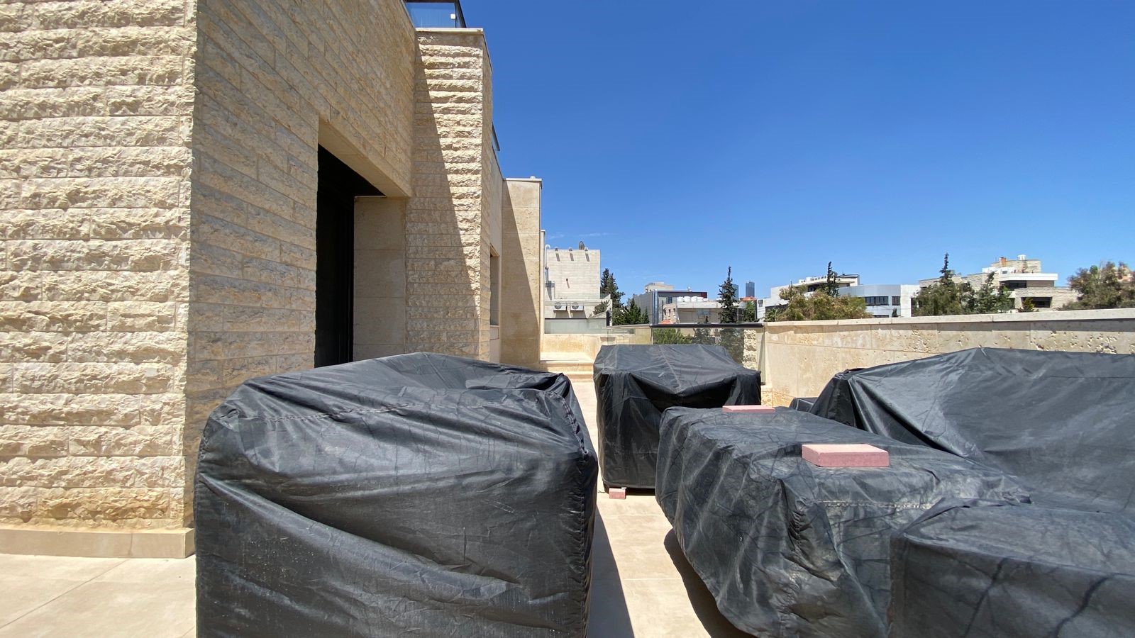 cfc945af-4aff-40da-9f5c-401af5ee6418 Rooftop terrace with beige stone walls and several large items covered in black tarps, against a clear blue sky.