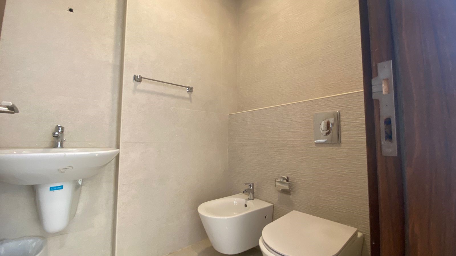 cffebc66-8c9f-4d4b-bb9d-721bd1660d74 Compact beige-tiled bathroom with a wall-mounted sink on the left, a bidet and a closed toilet along the right wall, and chrome fixtures.