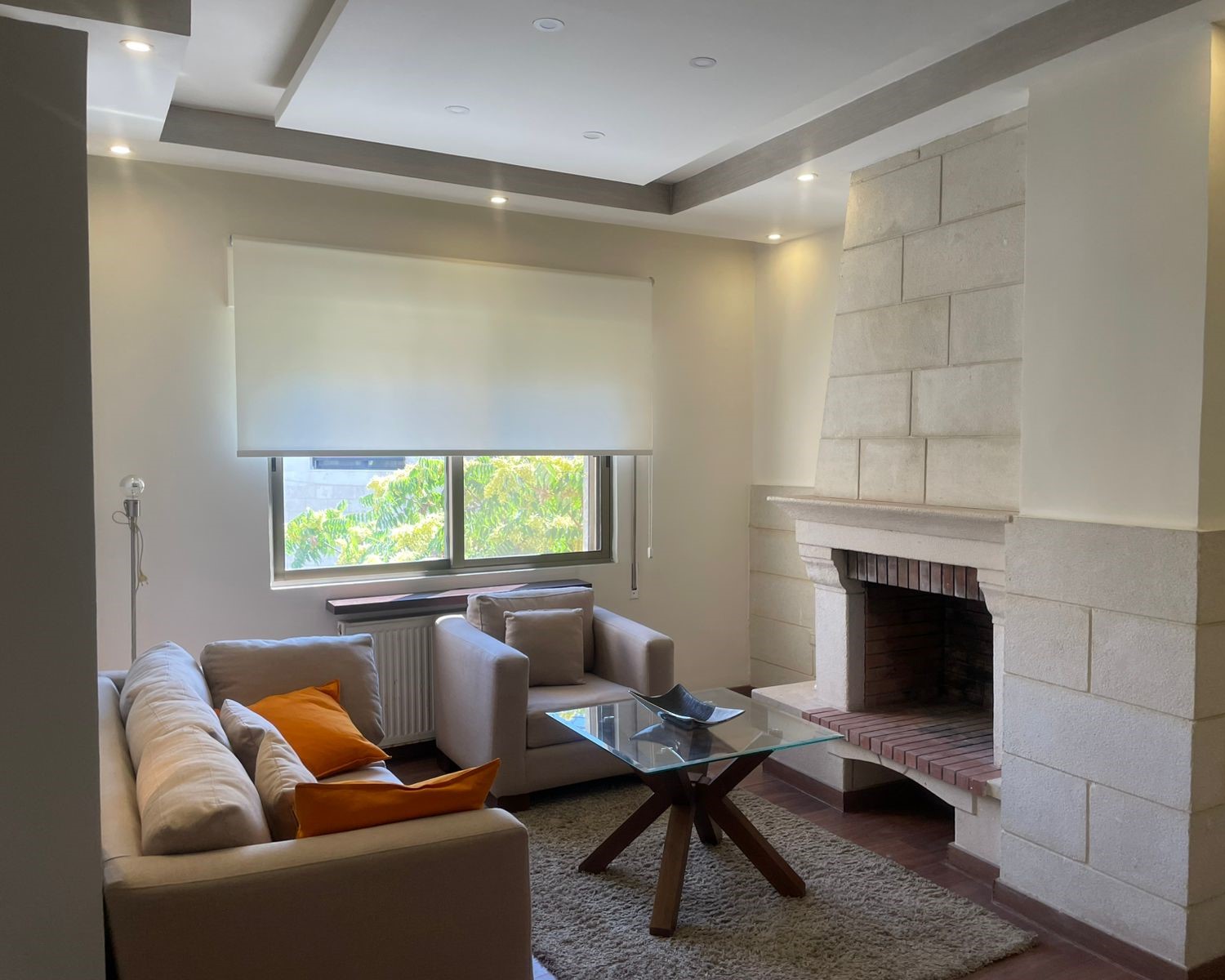 d27fbb50-74ce-4d2f-af53-fcddb45ff652 Modern living room with a stone fireplace, beige sofa, and orange cushions facing a glass coffee table.