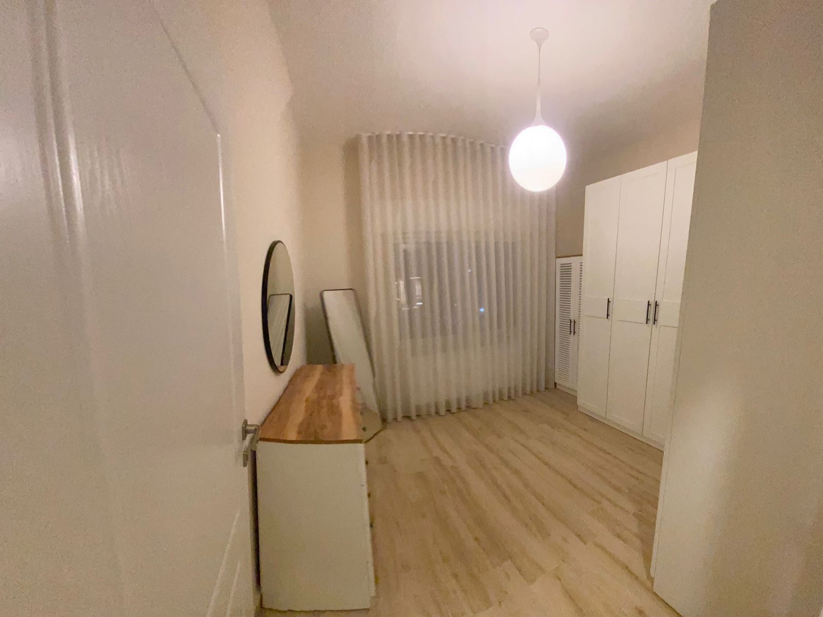 d338e090-fcad-4969-8ce0-b0b4b9a0bfcf Minimalist bedroom: white wardrobe, wooden dresser, round wall mirror, and a full-length mirror leaning by sheer curtains under a globe ceiling light.