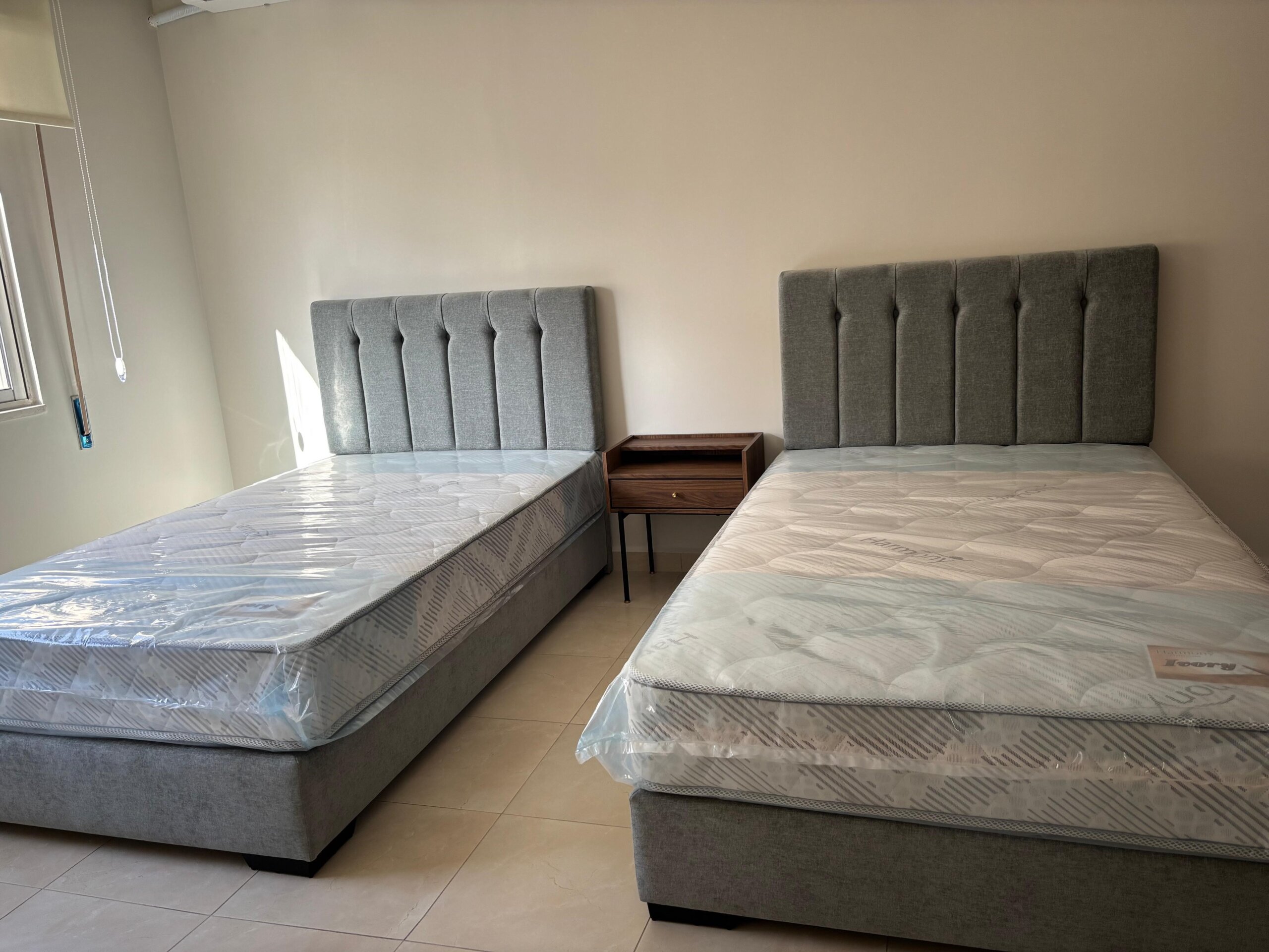 d3d61e82-cd72-47f6-a292-cd5313239617 Two twin beds with matching gray tufted headboards and a small wooden nightstand between them in a bright, minimal bedroom setting.