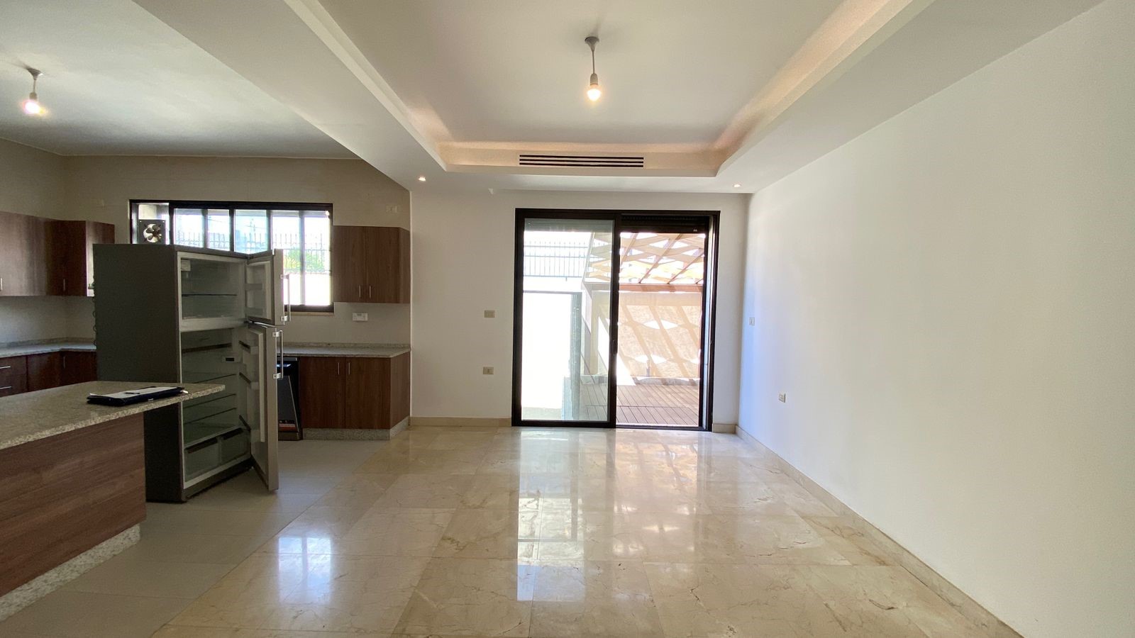 d3e895b6-4135-4b28-9225-d58d82201712 Open-plan kitchen and living area with marble floors, wooden cabinets, and a sliding glass door to an outdoor patio; fridge doors are ajar.