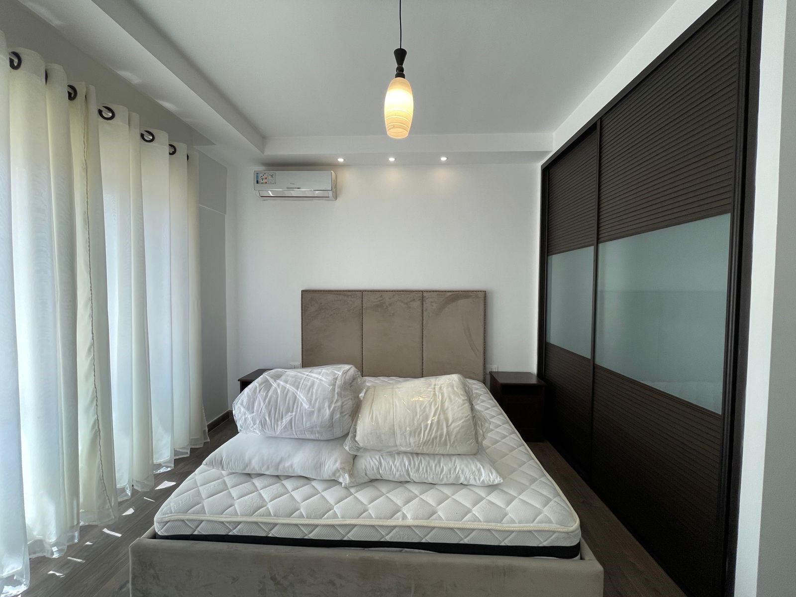 d433c232-3b58-4253-a518-a165268bba22 Modern bedroom with a white bed, stacked pillows, tufted headboard, and sheer white curtains on the left.