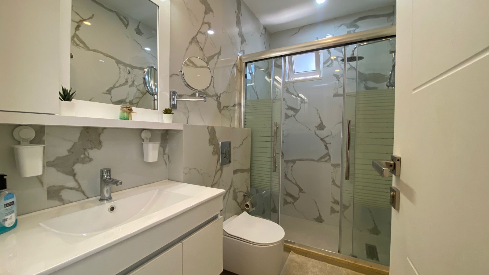 d493ca71-3052-4647-b615-a8f6fb307acb Modern bathroom with a marble-pattern wall, white vanity, and glass sliding shower doors propped by chrome fixtures.