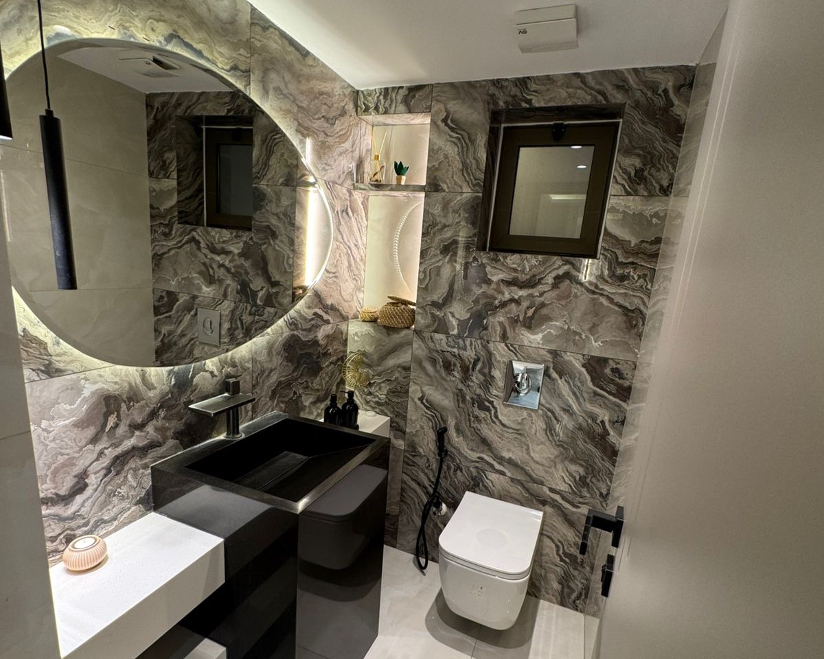 d527fbd8-b22f-4e68-afe9-36bdb8bc3a4c Modern bathroom with dark marble walls, a large round backlit mirror, and a black rectangular sink beside a wall-mounted toilet.