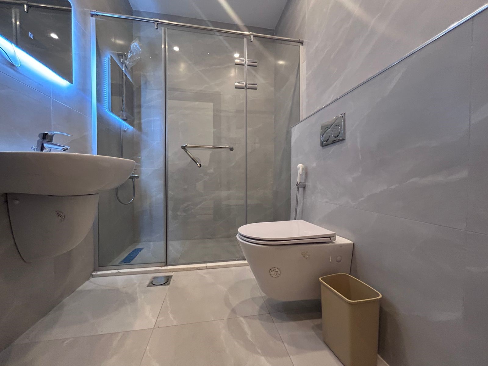 d5584520-c439-4b83-9768-0affd1e2948c Modern grey-tiled bathroom with a glass shower enclosure, wall-mounted sink, and a wall-mounted toilet with a beige trash bin nearby.