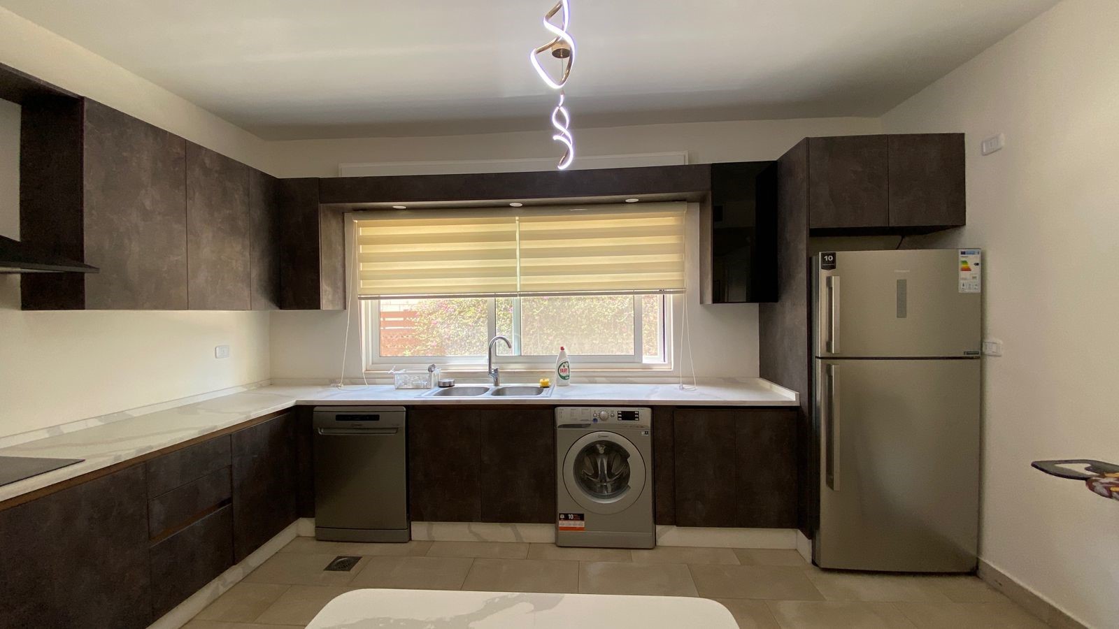 d5ba01e0-0ec0-4694-803d-fbbb07c98704 Modern L-shaped kitchen with dark wood cabinets, stainless steel appliances, and a window above the sink. A spiral pendant light hangs from the ceiling.