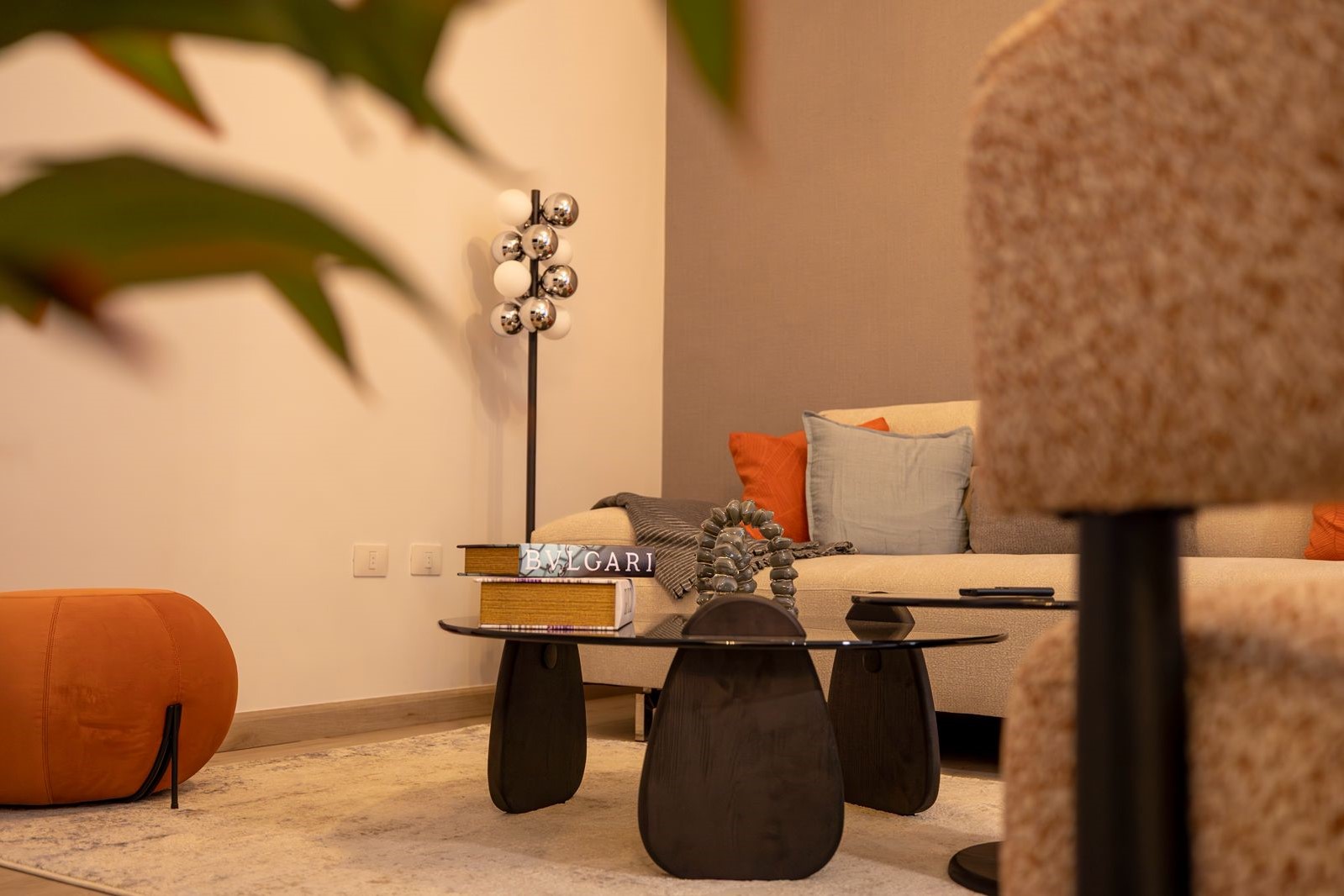 d5d50ac0-55a8-4f33-becf-475b2d30b5fe Cozy living room corner with a beige sofa, orange and pale blue cushions, a glass coffee table, a BVLGARI book, and decorative sculpture on display.
