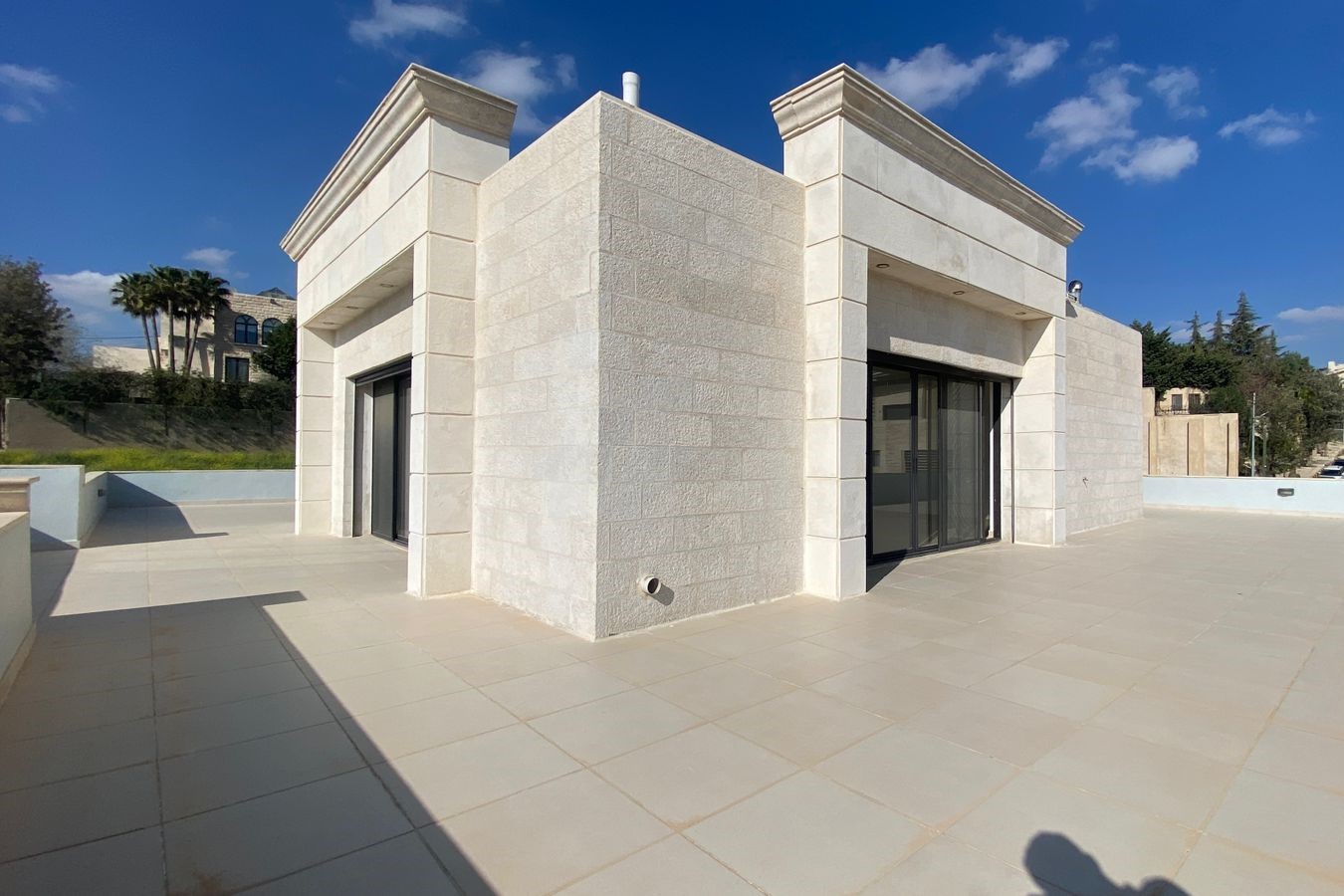 d62de3c8-b8b5-44b4-96a0-4d8a609dfee2 Modern beige stone house with large sliding glass doors on a wide tiled terrace under a blue sky.