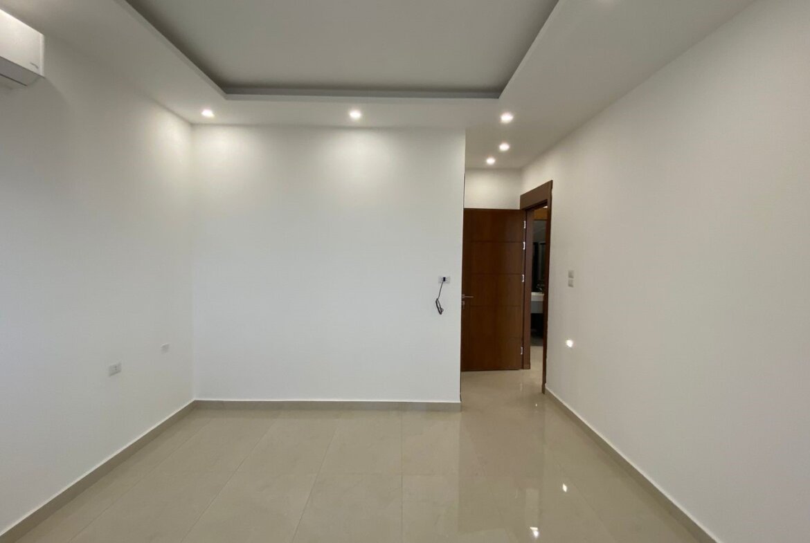 Empty white room with glossy beige floor tiles, recessed ceiling lights, and an open wooden door leading to another area; wall-mounted air conditioner on left.