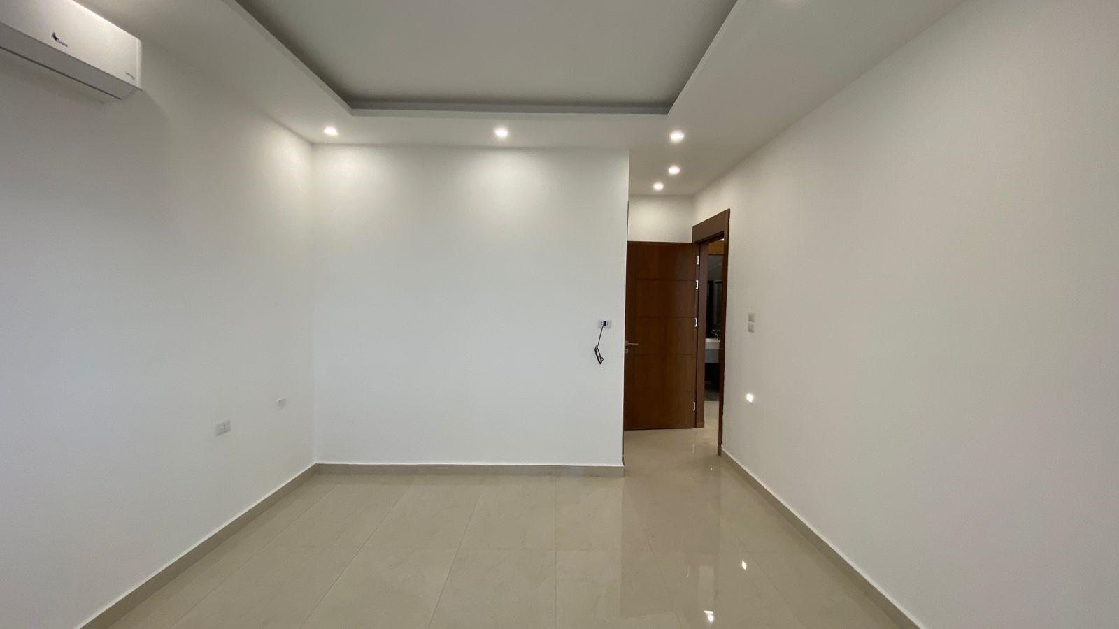 d6d9fbf4-7341-4b35-bb5c-7fec89403b70 Empty white room with glossy beige floor tiles, recessed ceiling lights, and an open wooden door leading to another area; wall-mounted air conditioner on left.