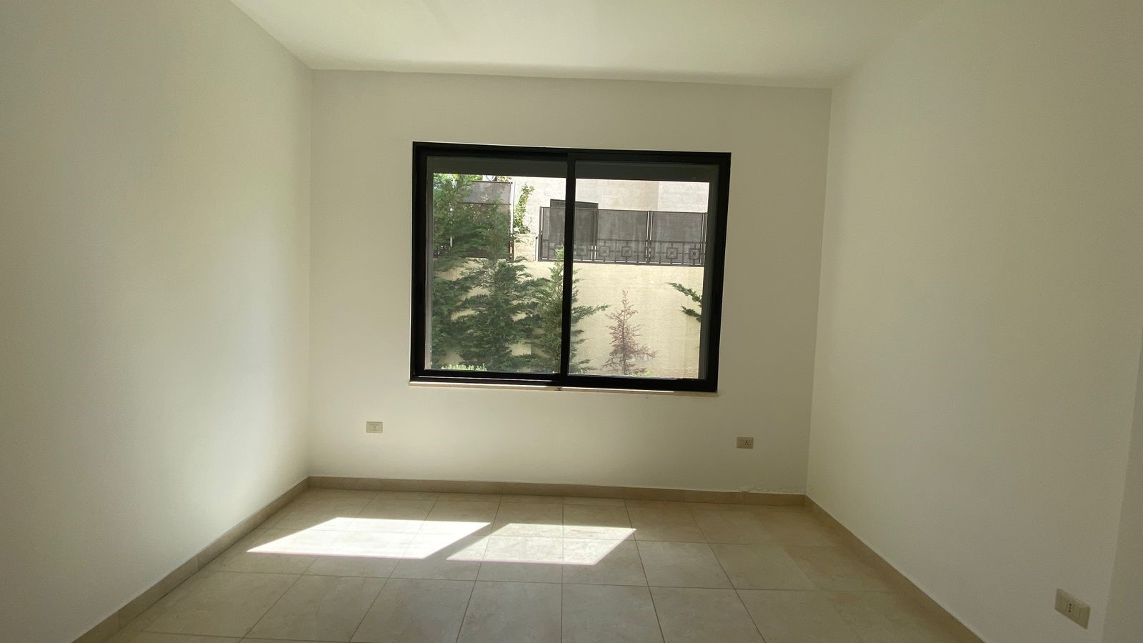 d8188426-c9ce-4b50-a7c9-9b8500f938bd Empty white room with a large black-framed window and sunlight on beige tiled floor.