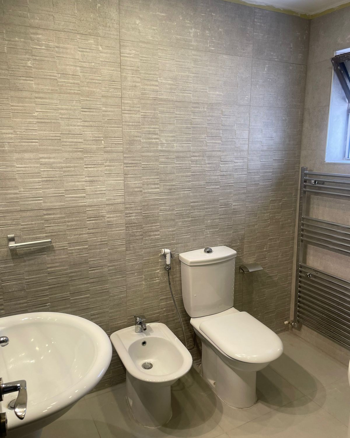 d850bd10-8697-4a99-b16e-255b1a67167b Modern bathroom with a white toilet, bidet, and sink arranged along a tiled wall, plus a chrome heated towel rail on the right.