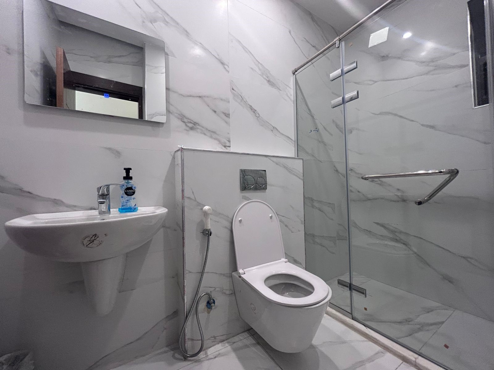 d858e015-da24-4d5b-936e-bff383458333 Marble-tiled bathroom with wall-hung sink, blue soap bottle, and a wall-mounted toilet adjacent to a glass shower enclosure.