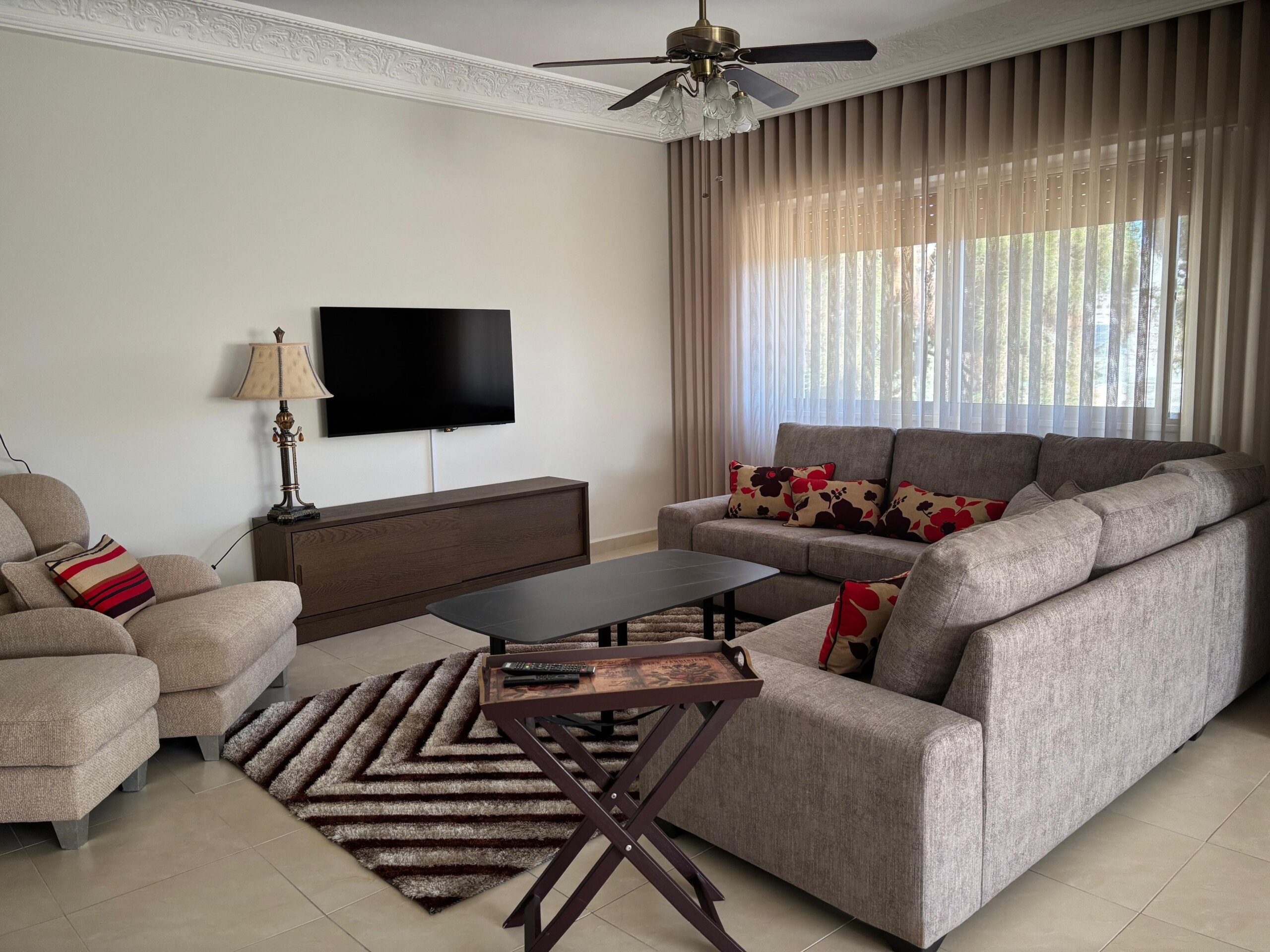 d97d646e-9b59-4365-b361-d9ee47b5451d Living room with a gray sectional sofa, patterned red pillows, two beige armchairs, a dark wood media console, and a wall-mounted TV under sheer window curtains.