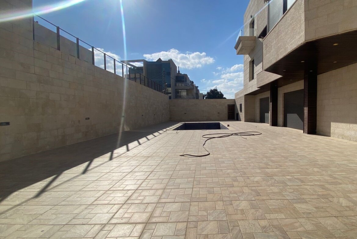 Sunny open courtyard with beige stone walls, tiled ground, a rectangular pool, and a coiled hose.