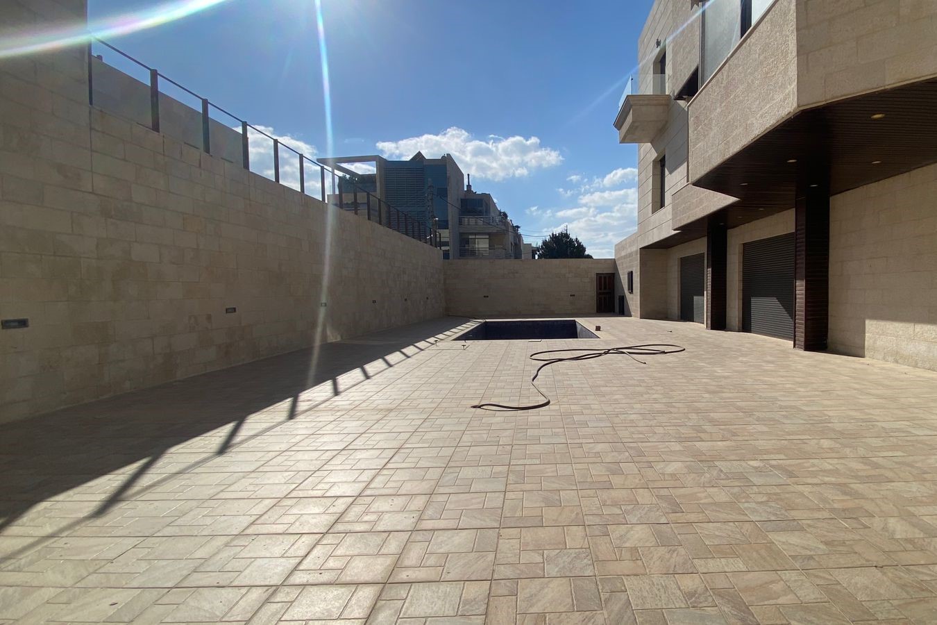 da098019-2e3a-4d7d-89ad-f27a318b0338 Sunny open courtyard with beige stone walls, tiled ground, a rectangular pool, and a coiled hose.