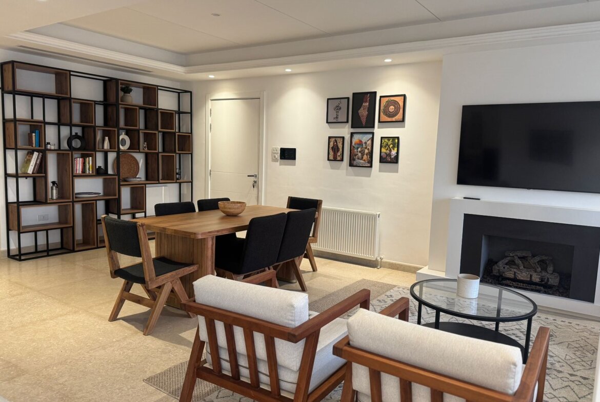 Modern living room with a wooden dining table and six black-cushioned chairs, a tall wood-and-metal bookshelf on the left, wall art on the right, and a fireplace with a TV above it.