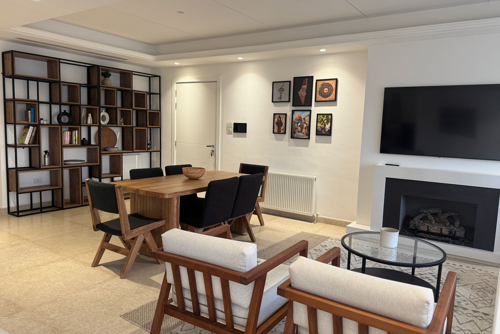 db7fdae1-17ba-4233-860c-10d9e1e8a08e Modern living room with a wooden dining table and six black-cushioned chairs, a tall wood-and-metal bookshelf on the left, wall art on the right, and a fireplace with a TV above it.