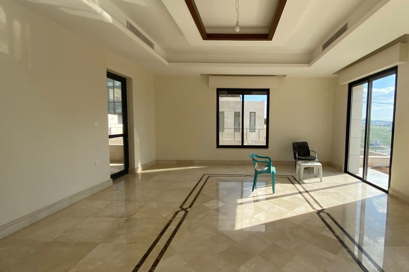 dc8654f2-8598-44c1-96d0-3e61e7569731 Bright, empty living room with beige marble floor, a decorative inlaid border, large windows, and a skylight; two plastic chairs and a small stack of stools near the corner.