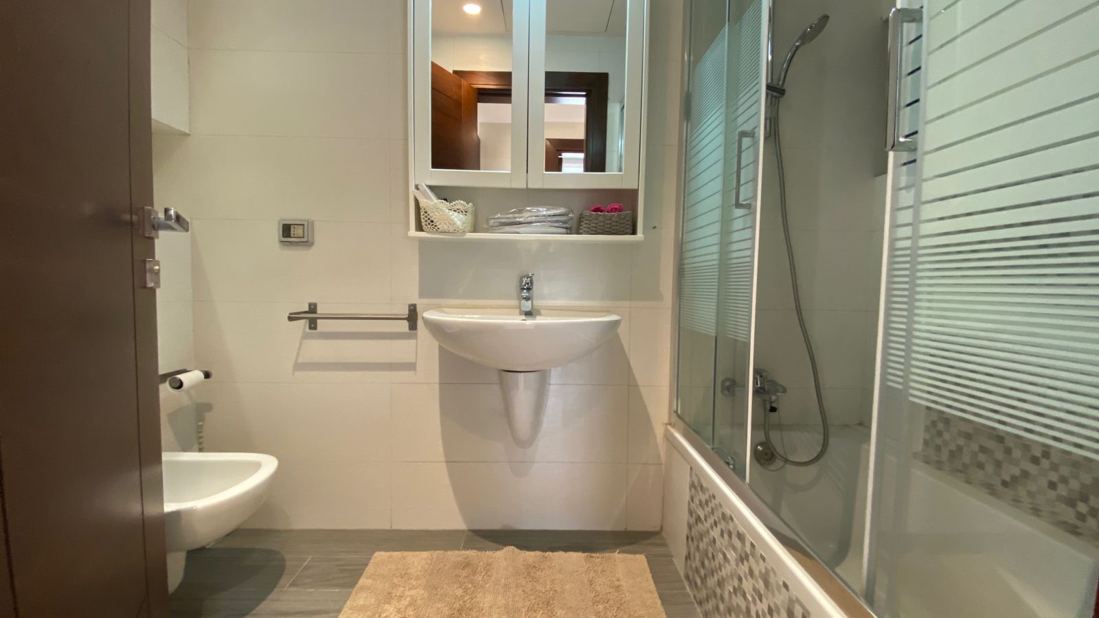 dde88273-5c61-437a-a690-3751a6ad4a5b Modern bathroom with white sink, wall mirror cabinet, towel rail, and glass-enclosed shower to the right; beige bath mat on gray tile floor.