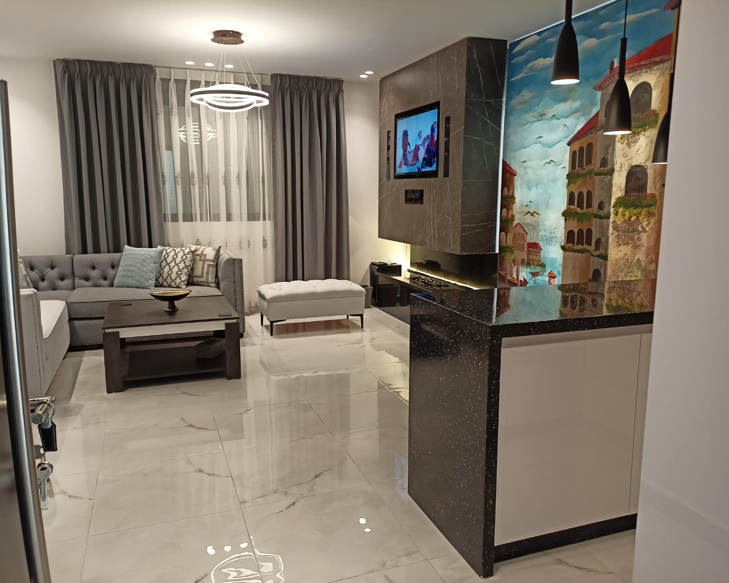 df5ac4ee-85fb-4766-8917-78d063e4d362 Modern living room with a gray tufted sectional, coffee table, and a wall-mounted TV set into dark marble panels; a colorful city mural occupies the right wall.