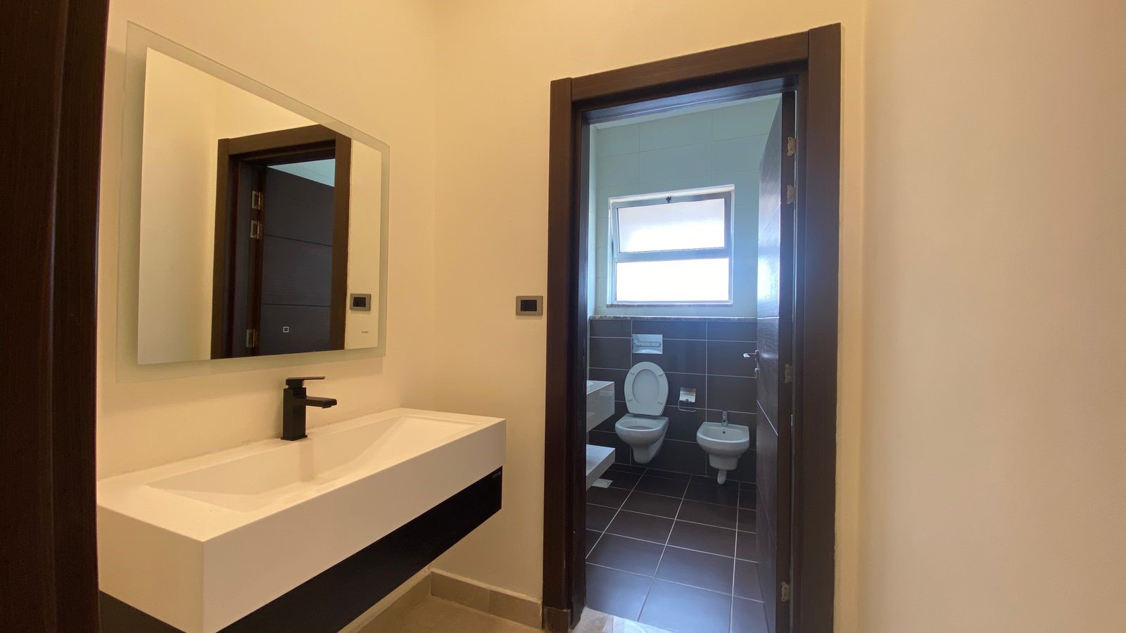 e0e74e40-8241-4733-b3cd-c263b08425bc Modern bathroom with a white rectangular sink and black faucet in the foreground; a doorway opens to a toilet and bidet area with dark floor tiles and a window.