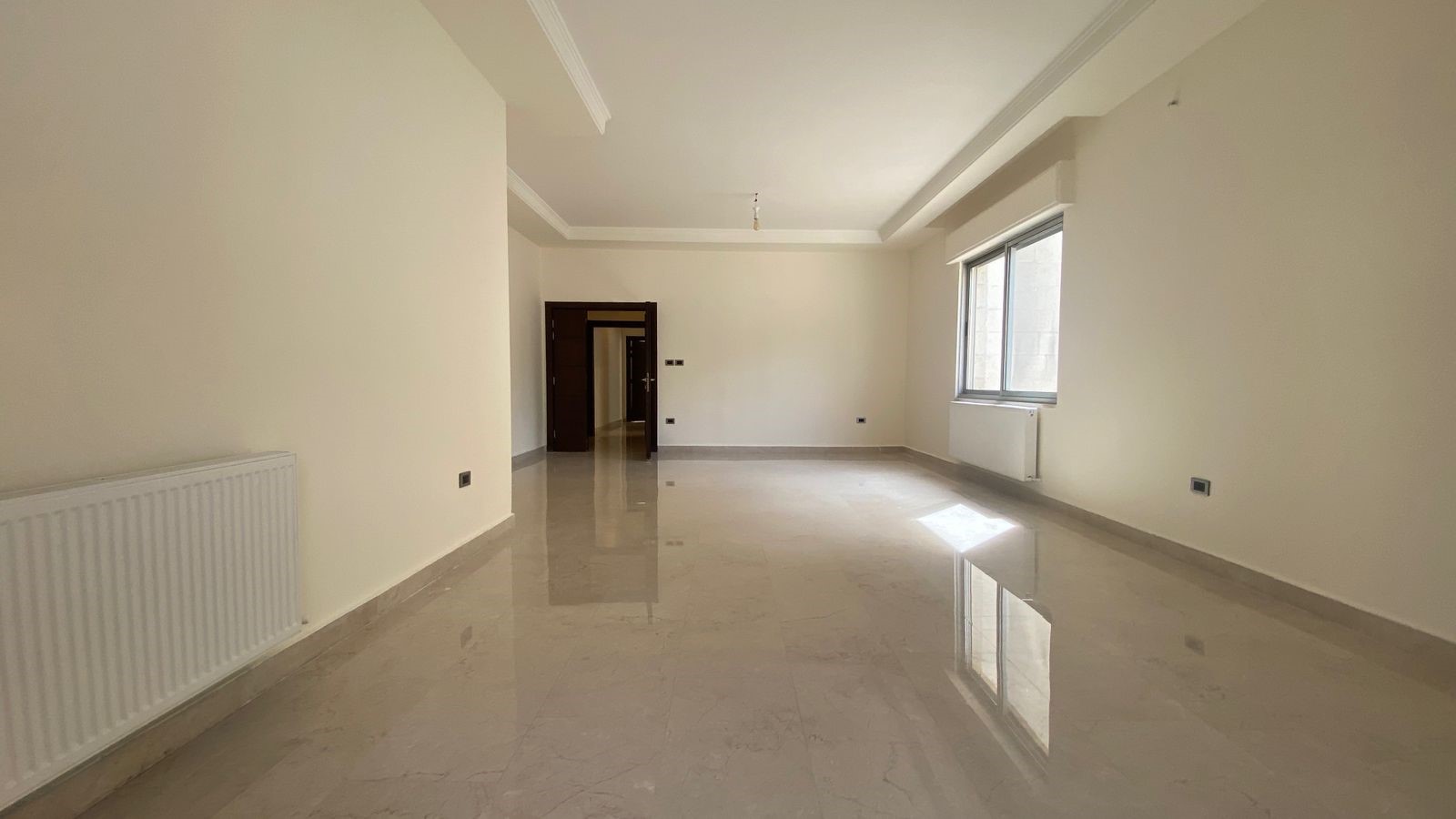 e2ff2fc1-af26-47ad-93f7-ee360dbcf4dc Spacious empty living room with white walls, glossy marble floor, radiator on the left, and a window on the right; doorway at the far end.
