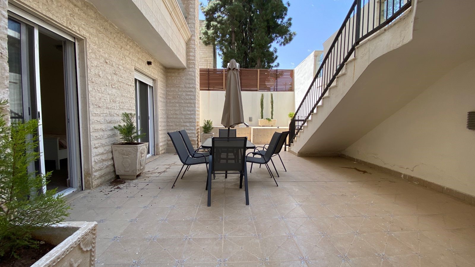 e4f2313a-e9d8-428d-9669-d797104e1e33 Outdoor patio with a six-chair dining set and a closed umbrella, flanked by sliding doors and a stairwell.