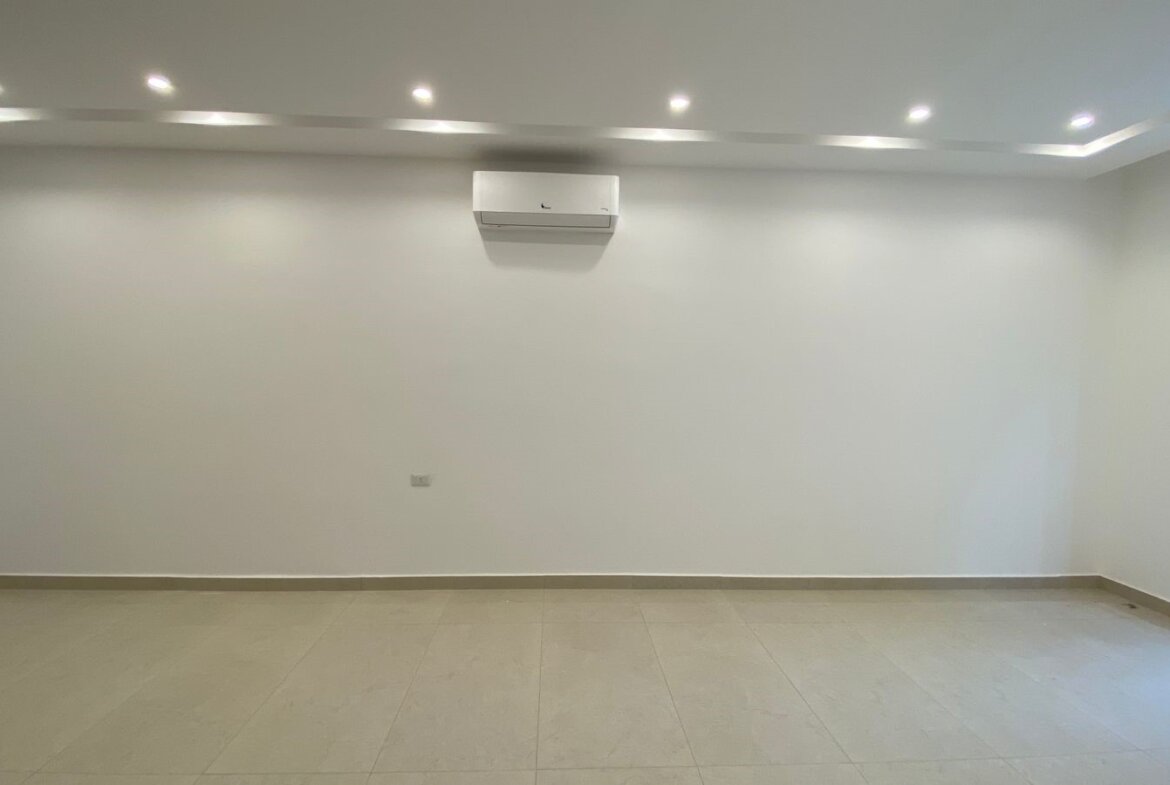 Empty white-walled room with tiled floor, recessed ceiling lights, and a wall-mounted air conditioner centered on the back wall.