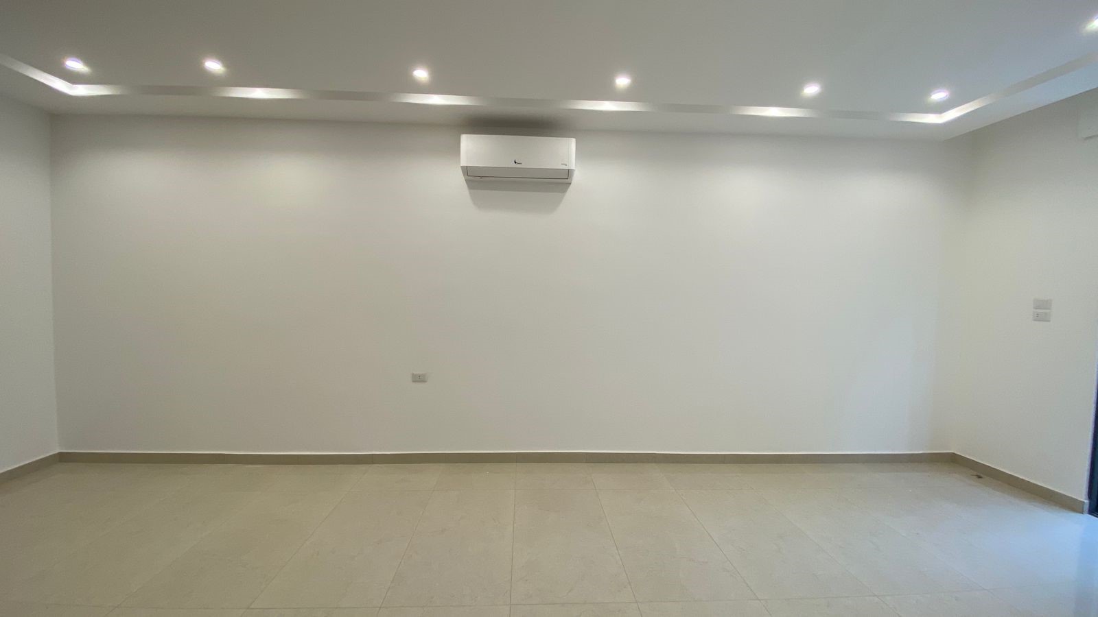e549232e-a243-4ca8-b2f1-5e456e0dccb2 Empty white-walled room with tiled floor, recessed ceiling lights, and a wall-mounted air conditioner centered on the back wall.