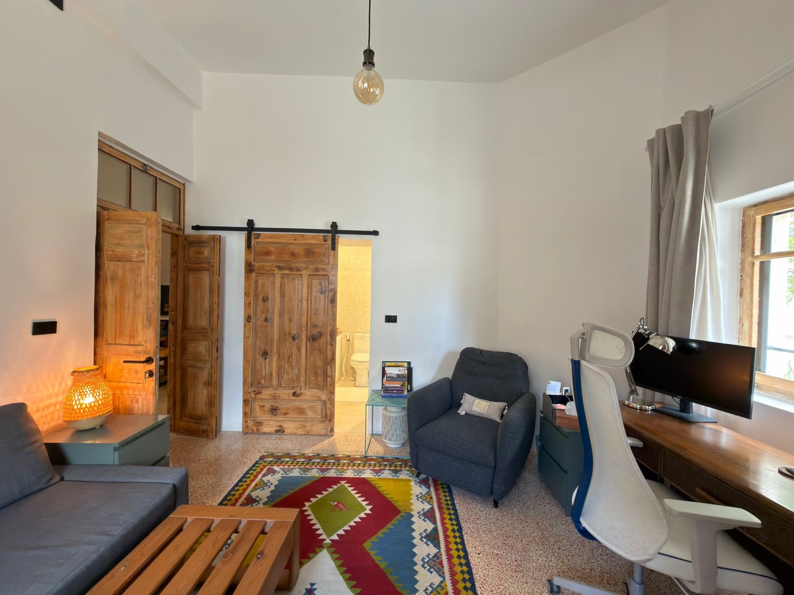 e6441901-36d1-4358-bede-a1d7a31d4da2 Bright living space with rustic wooden doors, a gray sofa, blue-green side table, and a corner desk with a large monitor by a window. A colorful geometric rug anchors the room.