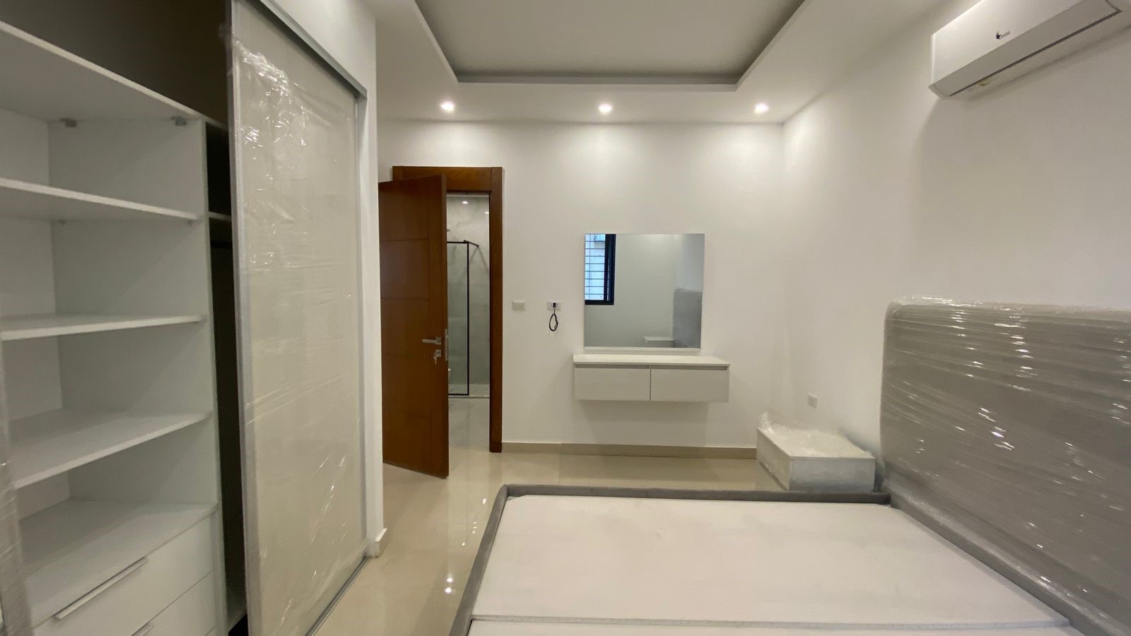 e6bec036-e018-4367-a4ca-b0a74aa4cead Modern bedroom scene with a wrapped mattress, white shelving, and a sliding-door wardrobe; open wooden door to an en-suite area visible in the background.