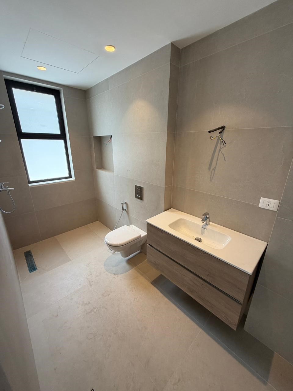 e7f771bf-34c1-4421-abe1-b5700303d353 Modern bathroom with a wall-hung toilet, white sink vanity, and large beige tile walls and floor; window on the left.