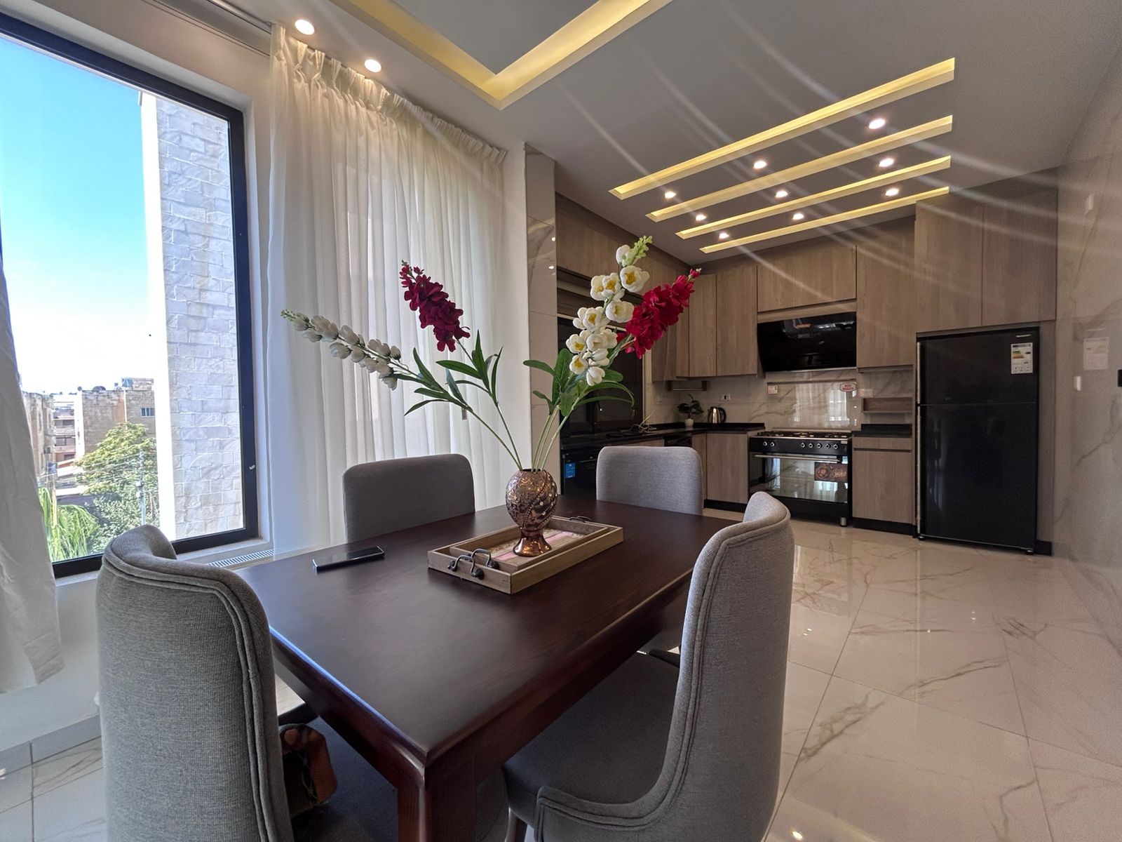 e97a42aa-af62-4eb2-b345-90fcfe732d30 Modern dining room with a dark wooden table, gray upholstered chairs, and a vase of red and white gladiolus; kitchen in the background.