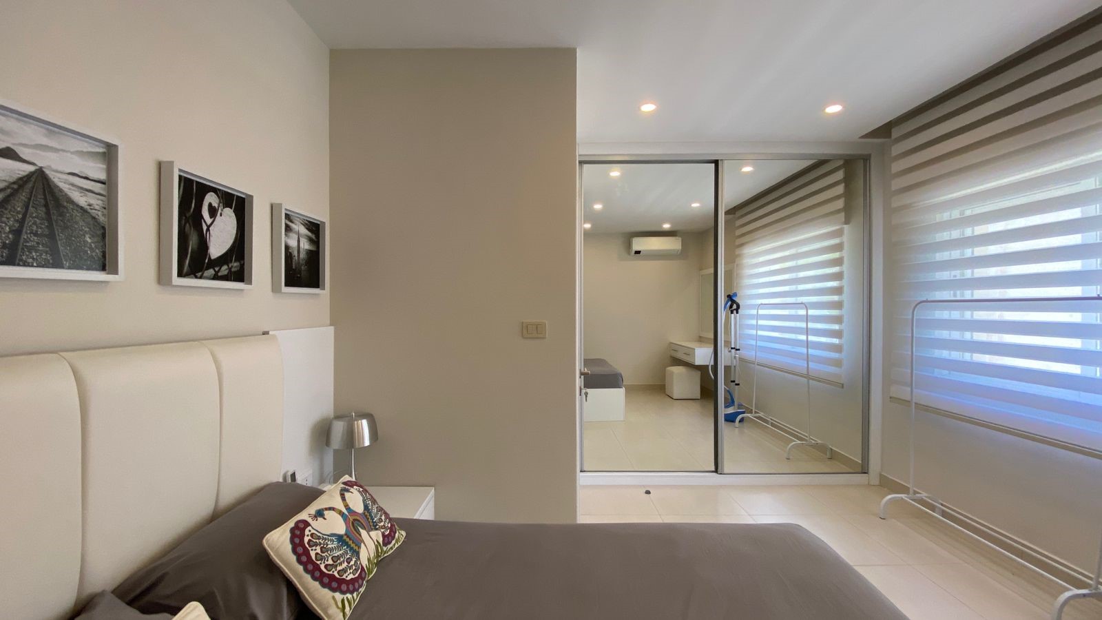 ea2bf340-d795-4a71-a3d7-d4b89a3d2b25 Modern bedroom with white upholstered headboard, gray bedding, and decorative pillows; glass sliding door opens to an adjoining area with blinds and a small workspace.