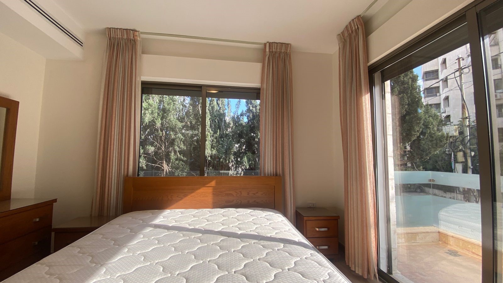 eb5d9978-9e52-426b-9ea7-ea19510002d4 Sunlit bedroom with a wooden bed, matching nightstands, and large windows with beige curtains showing trees outside.